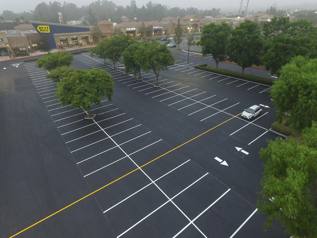 Parking lot of a Best Buy location with yellow exit sign, marked lanes, and trucks visible in the background under a clear blue sky.